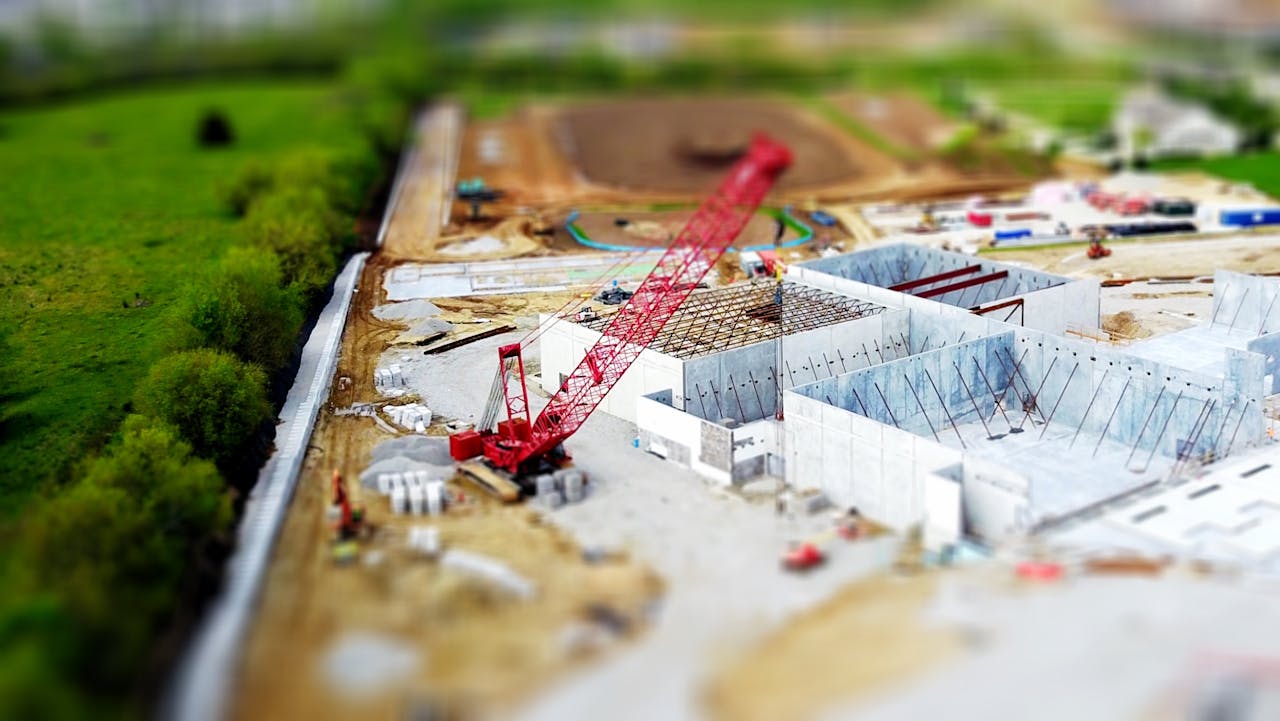 services-03 Aerial view of a construction site with a red crane amidst green fields and concrete buildings.